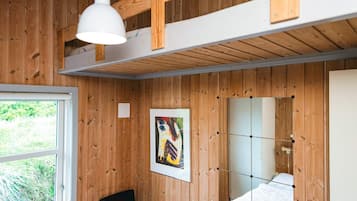 A room with wooden walls, a bed with a white mattress, a black chair, a white pendant light, and a window with a view of greenery.