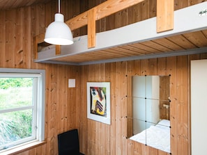 A room with wooden walls, a bed with a white mattress, a black chair, a white pendant light, and a window with a view of greenery.