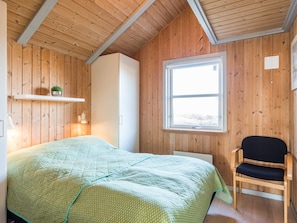 A room with wooden walls, a bed with a green quilt, a chair, a window, and a shelf with a plant. - 4 Star Holiday Home in Henne (Henne)