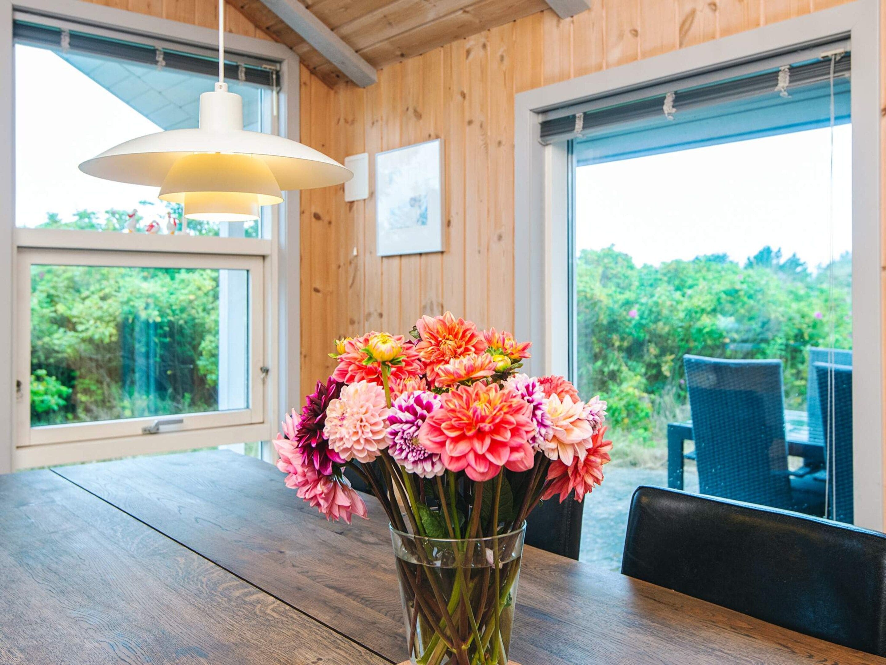 A wooden interior with a dining table, a vase of flowers, and a view of greenery through large windows.