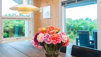 A wooden interior with a dining table, a vase of flowers, and a view of greenery through large windows.