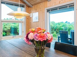 Interior de madera con una mesa de comedor, un jarrón de flores y vistas a la vegetación a través de amplias ventanas.