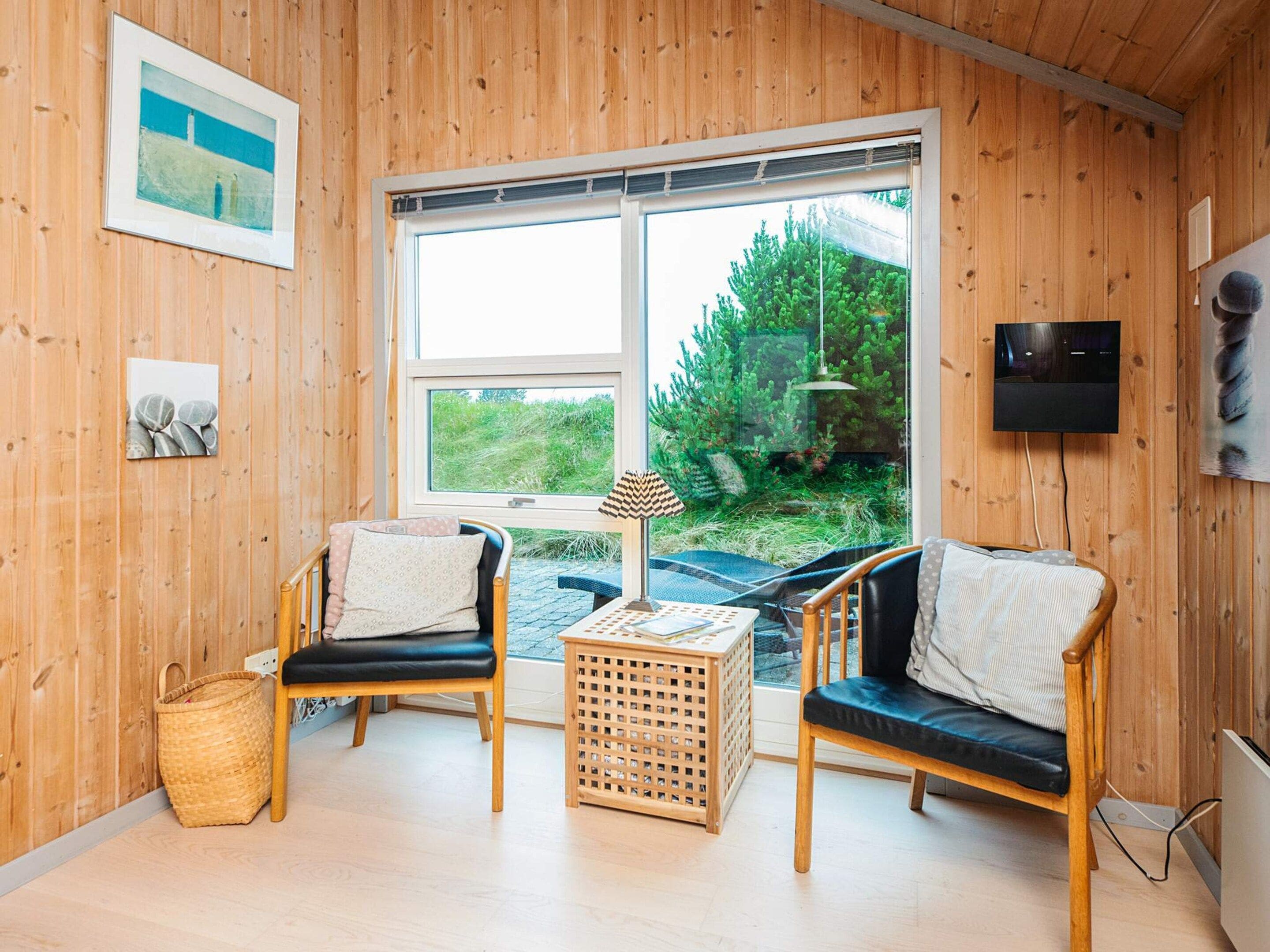 Een knus vertrek met houten wanden, twee zwarte leren fauteuils, een kleine houten tafel en een groot raam met uitzicht op groen.