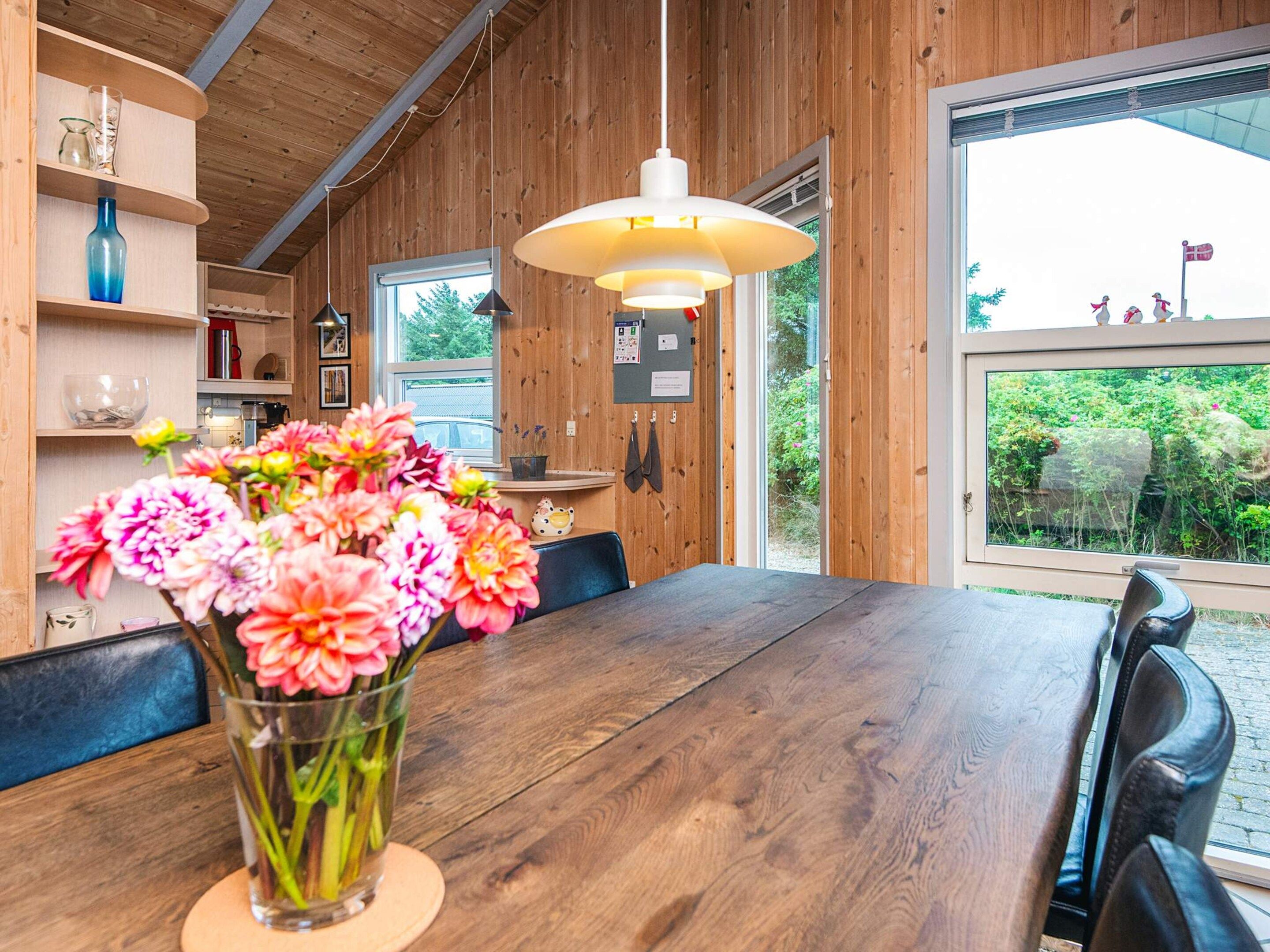 A wooden interior with a dining table, a vase of flowers, and a large window with a view of greenery.