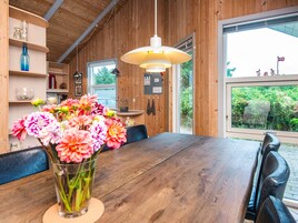 A wooden interior with a dining table, a vase of flowers, and a large window with a view of greenery.