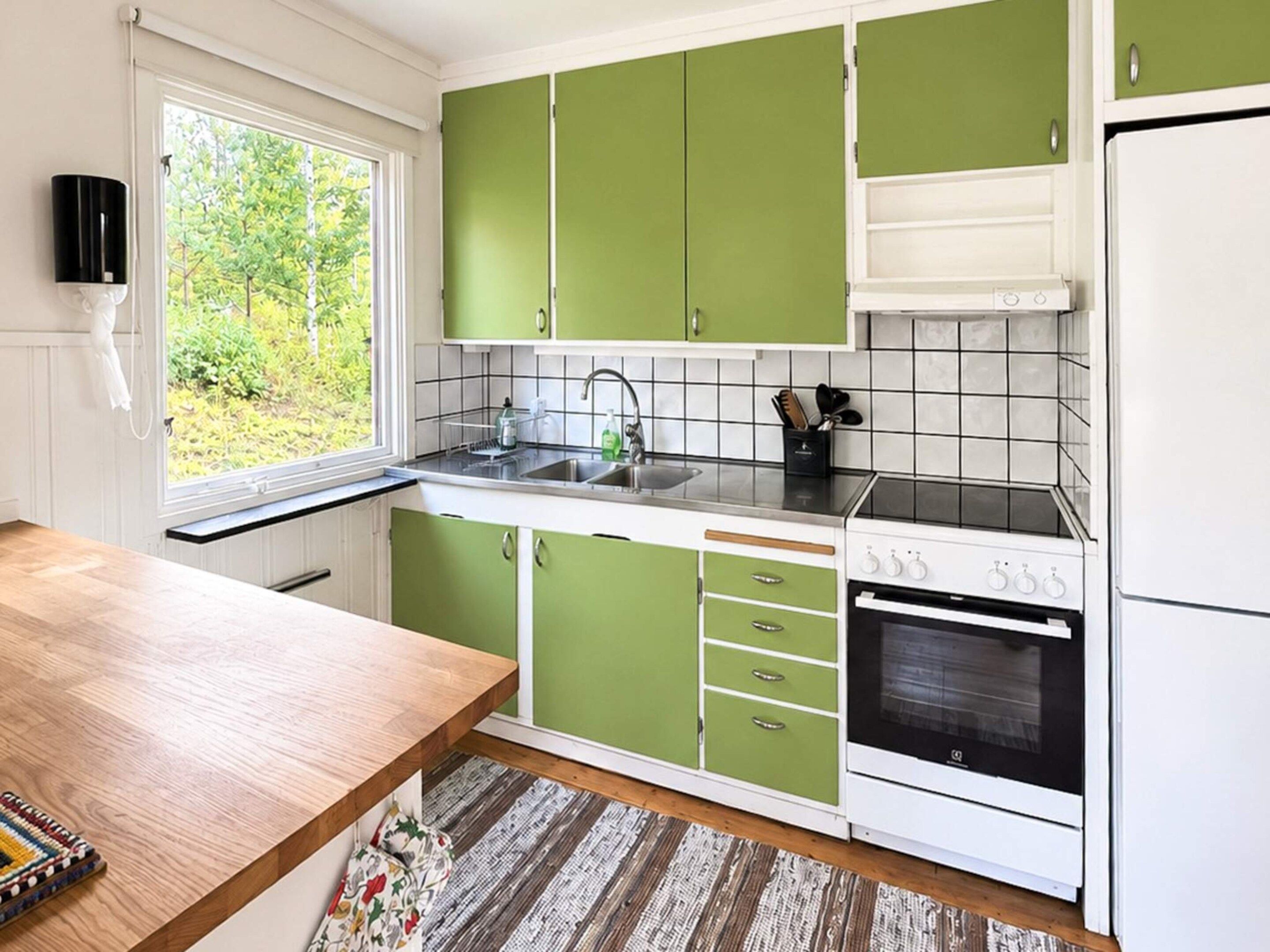 Une cuisine avec des meubles de couleur verte, un plan de travail blanc, un évier, une cuisinière et une fenêtre donnant sur un paysage verdoyant.