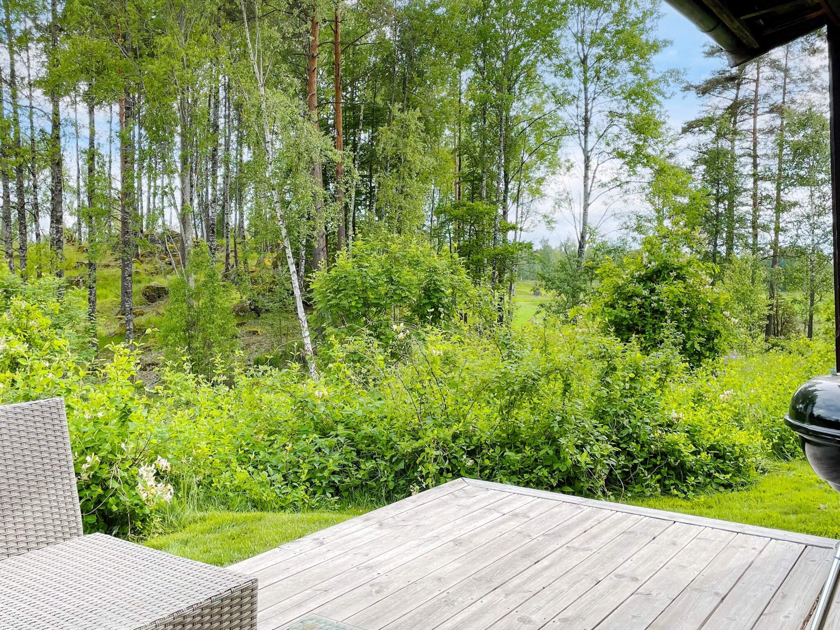 Une terrasse en bois, meublée de meubles en osier, entourée d’une végétation luxuriante et de grands arbres.
