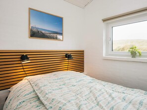 A bedroom with a wooden headboard, a bed with a patterned blanket, a nightstand with a lamp, a window with a view of the beach, and a framed picture on the wall. - 4 Star Holiday Home in Hvide Sande (Hvide Sande)