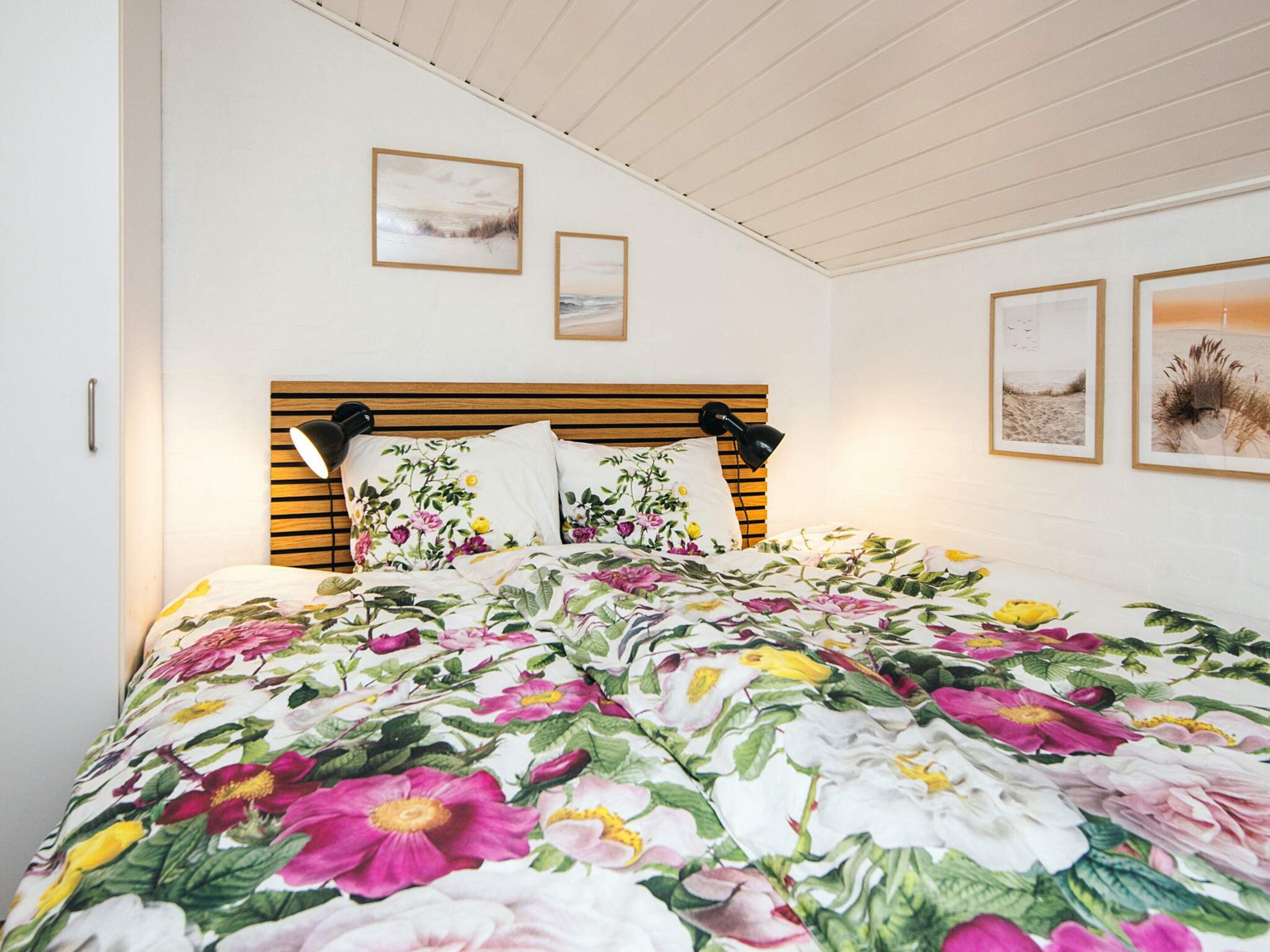A bedroom with a wooden headboard, floral bedding, two wall-mounted lamps, and framed artwork.