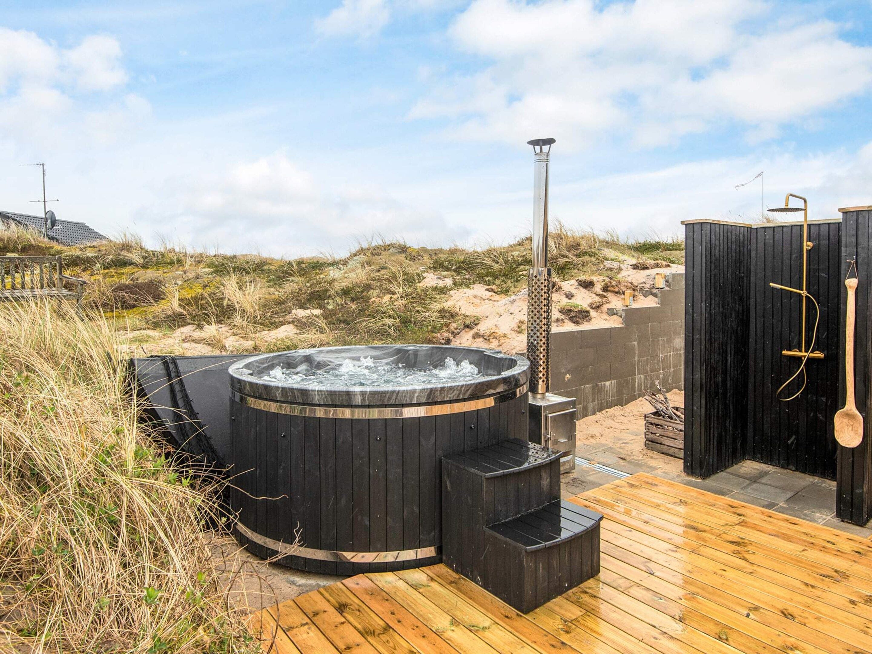 Um deck de madeira com hidromassagem e área de sauna, rodeado por dunas naturais e vegetação.