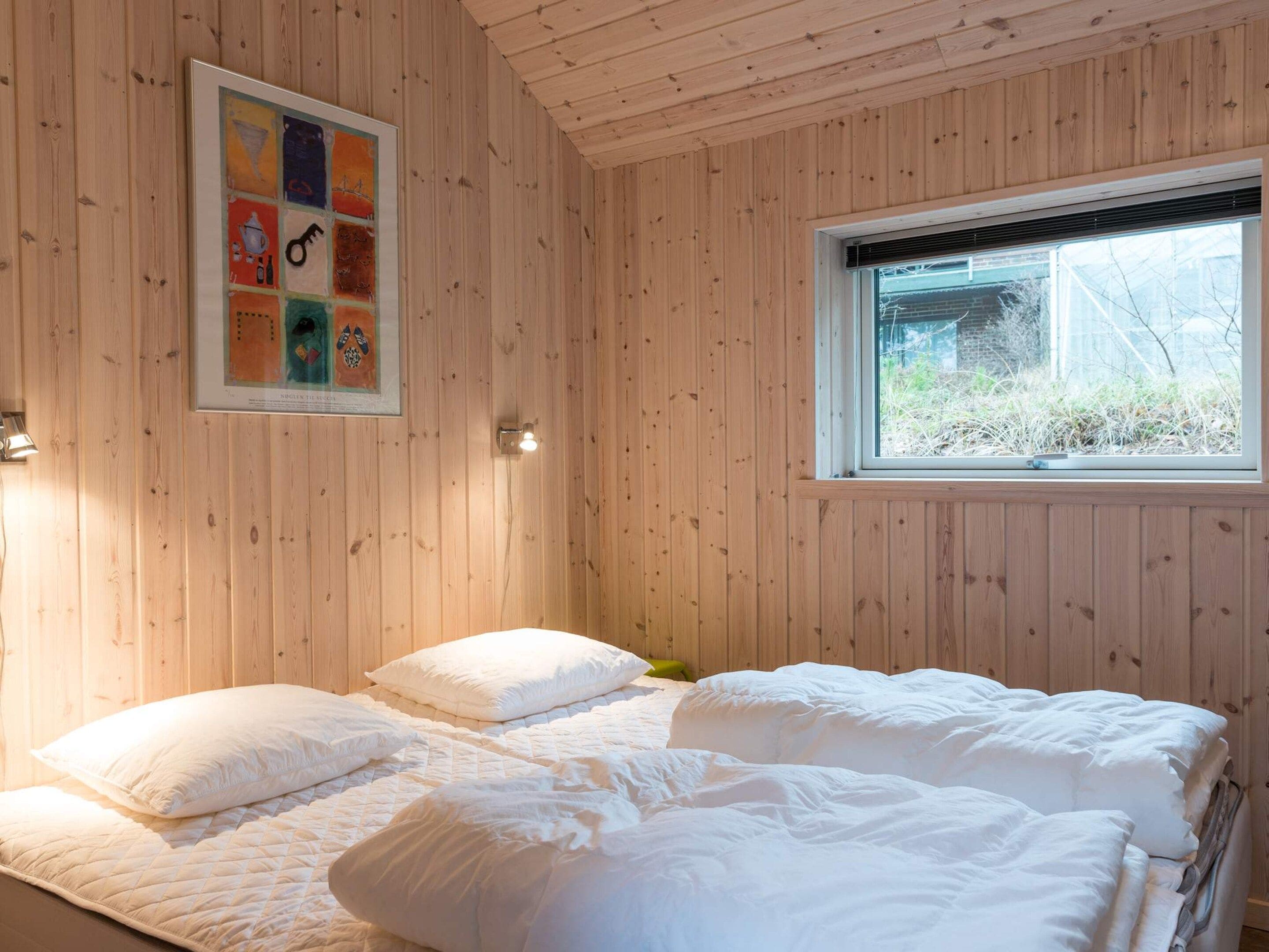 A room with wooden walls, a bed with white bedding, a window with blinds, and a framed picture on the wall.