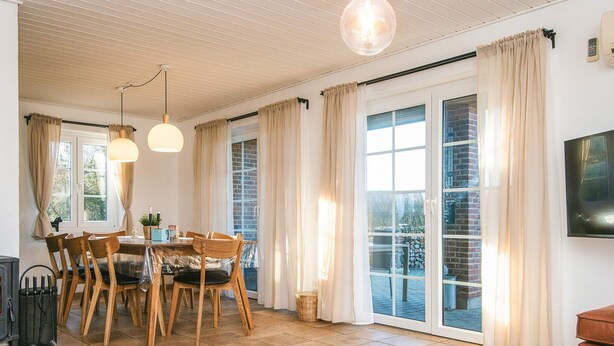 Uma sala de estar ampla com uma mesa de jantar, cadeiras e lareira. Há janelas grandes com cortinas, uma televisão de tela plana e um lustre.