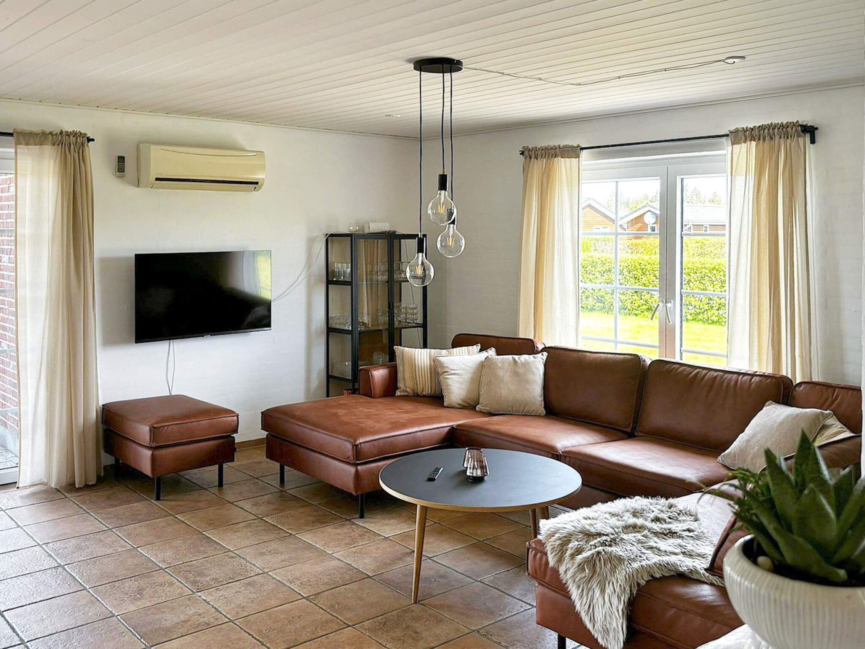 A living room with a brown leather sofa, a small round coffee table, a television mounted on the wall, and a potted plant.