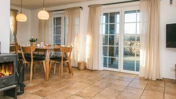 Une pièce chaleureuse avec une cheminée, une table à manger avec des chaises, un téléviseur fixé au mur et de grandes fenêtres avec des rideaux.