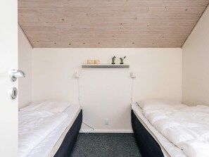 A small, single-room accommodation with two single beds, a wooden ceiling, a white wall, a shelf with decorative items, and a power outlet on the wall. - 4 Star Holiday Home in Henne (Henne)
