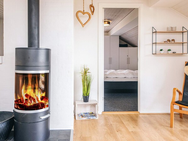 A modern living room with a wood-burning stove, a bed in the adjacent room, and a small plant on a side table.