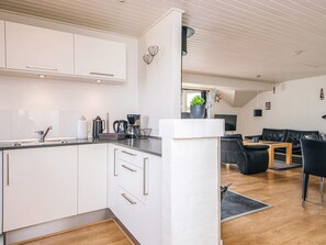 A modern kitchen with white cabinets and a wooden floor, leading to a living area with a black leather sofa and a coffee table. - 4 Star Holiday Home in Henne (Henne)