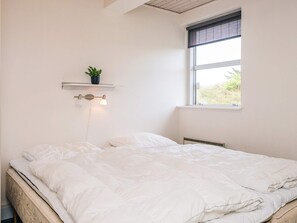 A neatly made bed with white bedding, a small potted plant on a shelf above, and a window with a view of greenery outside.