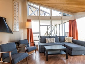 A modern living room with a sofa set, a coffee table, and a lamp. There is a large window with curtains and a view of a staircase.