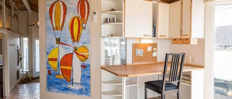 A modern kitchen with white cabinets, a wooden dining table, and a chair. There is a corkboard with notes and a painting of hot air balloons on the wall.