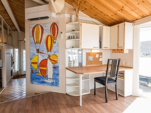 Une cuisine moderne avec des meubles blancs, une table à manger en bois et une chaise. On y trouve un tableau d’affichage en liège où sont accrochées des notes, ainsi qu’un tableau représentant des montgolfières.