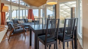 Interior moderno com teto de madeira, janelas amplas, área de jantar com cadeiras e mesa, e um sofá com almofadas.