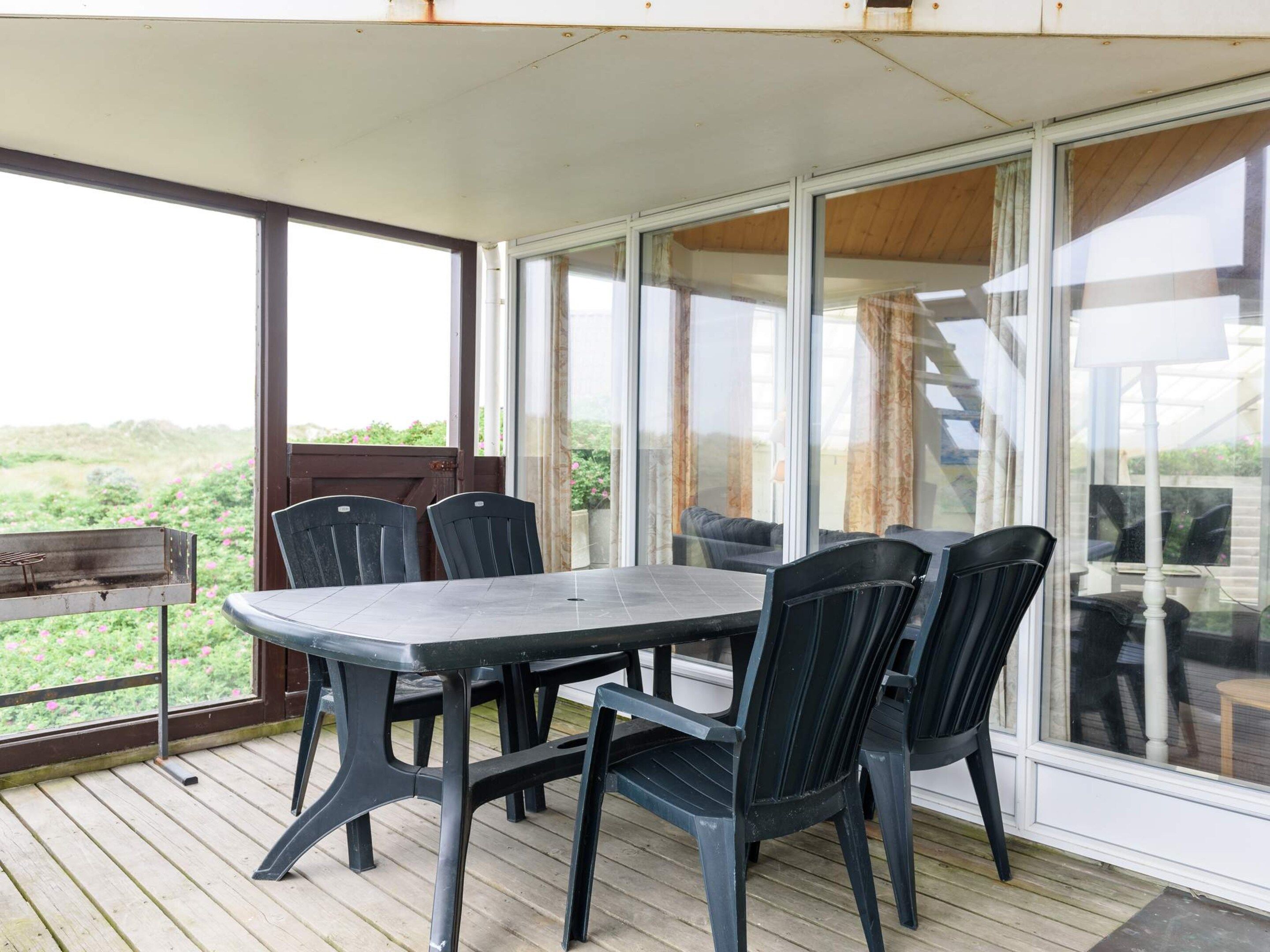 A covered outdoor dining area with a table and chairs, large windows, and a view of greenery.