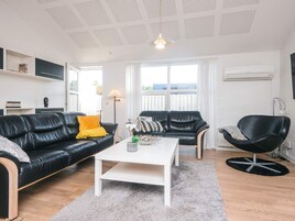 A modern living room with a black leather sofa, a white coffee table, and a black chair. There is a window with curtains, a floor lamp, and a ceiling light fixture.