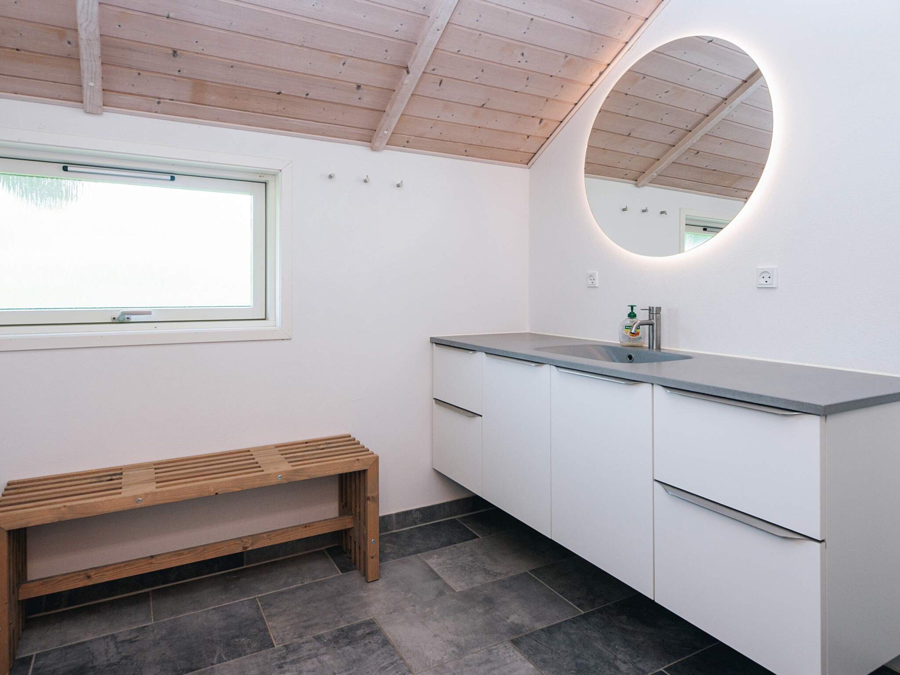 A bathroom with a wooden bench, a sink with a mirror, and a window.