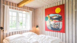 A bedroom with a wooden ceiling, a bed with white bedding, a window with curtains, and a colorful painting on the wall. - 4 Person Holiday Home in Norre Nebel (Norre Nebel)