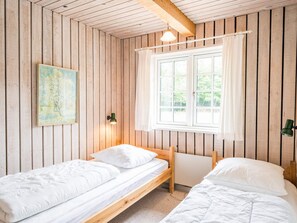 Une pièce avec deux lits en bois, une fenêtre avec des rideaux, une lampe fixée au mur et un tableau.