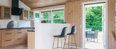 A modern kitchen with wooden cabinets, a central island with bar stools, and a view of the outdoors through large windows.