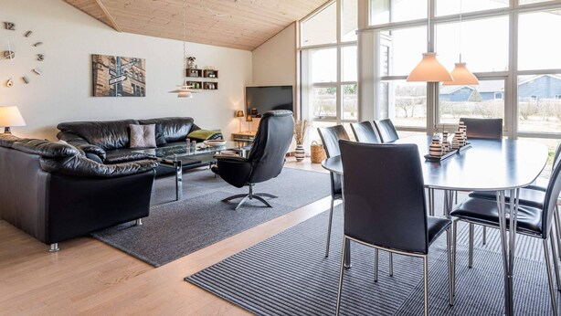 Una sala de estar moderna con un sofá de cuero negro, una mesa de centro de vidrio y un comedor con una mesa redonda y sillas. Amplias ventanas ofrecen luz natural y vistas al exterior.