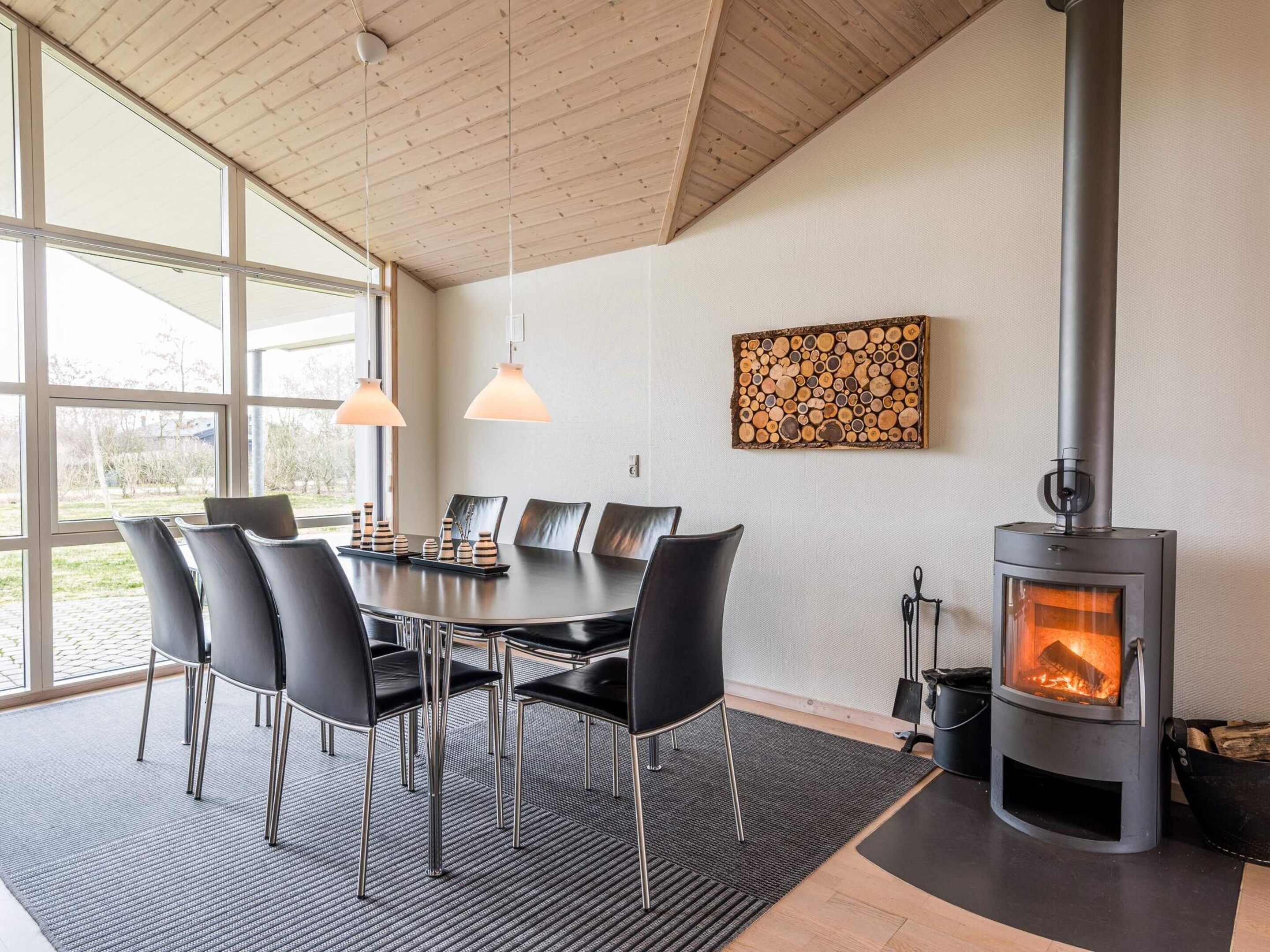 A modern dining area with a large table, chairs, pendant lights, a wood-burning stove, and a view through large windows.