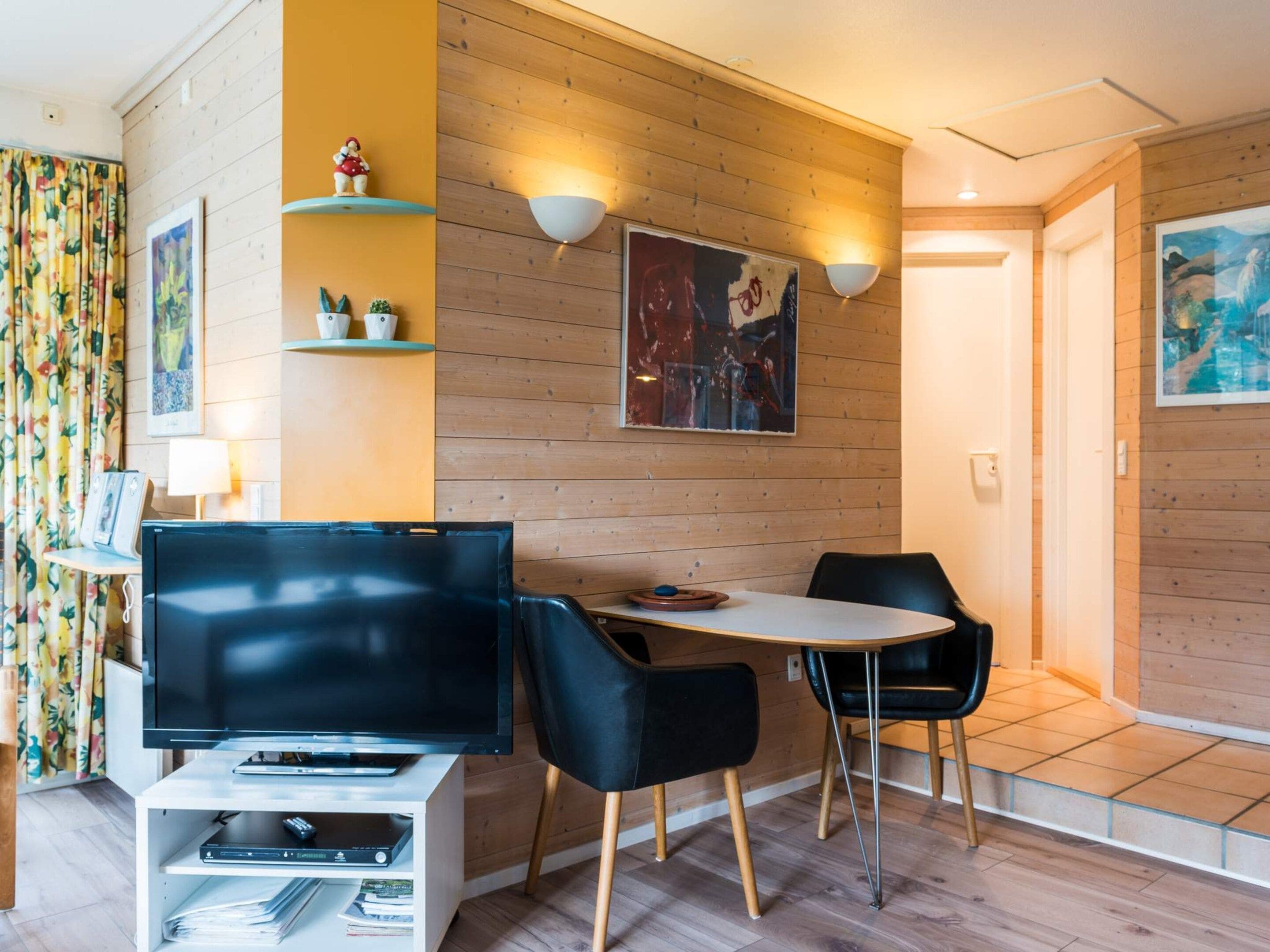 A modern living room with a flat-screen TV, two black chairs, a small table, wooden panel walls, and a painting.
