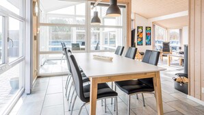A modern dining area with a large table and chairs, large windows, and wooden ceiling beams.