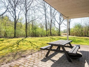 Et picnicbord under et halvtag med en flisebelagt terrasse, omgivet af træer og græs.
