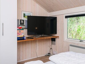 Une chambre avec un téléviseur fixé sur une étagère en bois, un lit avec une literie blanche, une fenêtre donnant sur un paysage verdoyant et un radiateur.