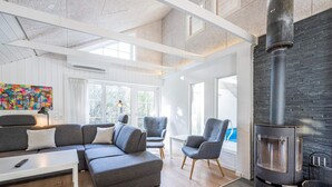 A modern living room with a grey sofa, two armchairs, a wooden floor, a large window, and a black wood-burning stove. - 4 Star Holiday Home in Blavand (Blavand)