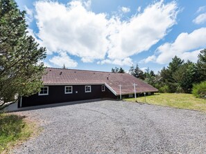A single-story building with a sloped roof, a gravel driveway, and a surrounding grassy area with trees. - 4 Star Holiday Home in Blavand (Blavand)
