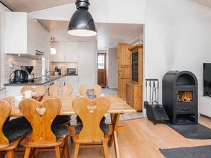 A modern kitchen with white cabinets, a wooden dining table with heart-shaped chairs, a black hanging light fixture, and a wood-burning stove. - 4 Person Holiday Home in Blavand (Blavand)