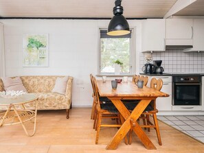 Ein gemütliches Wohnzimmer mit einer Couch, einem Esstisch mit Stühlen, einem Korbsessel, einer schwarzen Pendelleuchte und einer Küchenzeile mit Herd und Schränken.