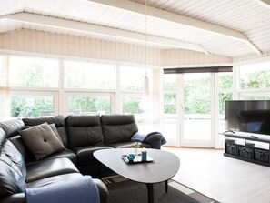 A modern living room with a leather sectional sofa, a coffee table, a flat-screen TV, and a large window offering a view of greenery.