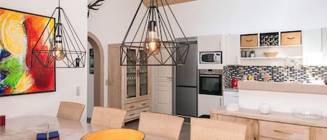 A modern kitchen with a dining area, featuring a large painting, geometric pendant lights, and a wooden ceiling.