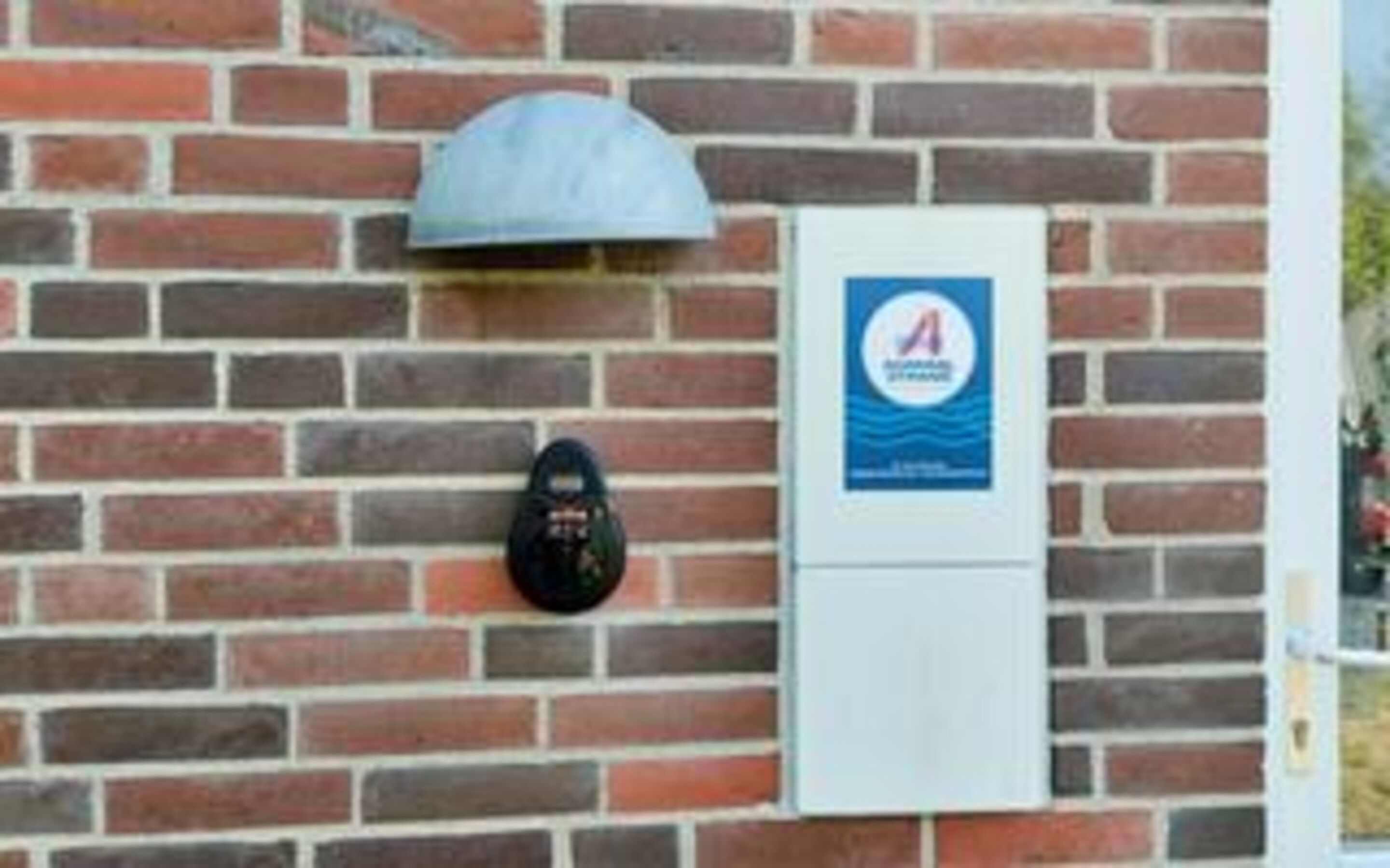 Un muro de ladrillo con un letrero azul y una caja blanca, posiblemente un medidor o panel de control.