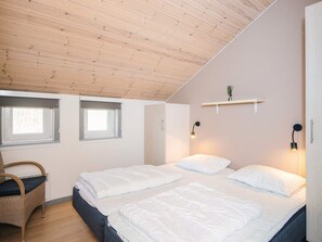 Une chambre avec un plafond en pente, deux fenêtres, un lit avec une literie blanche, une chaise en osier et des luminaires fixés au mur.