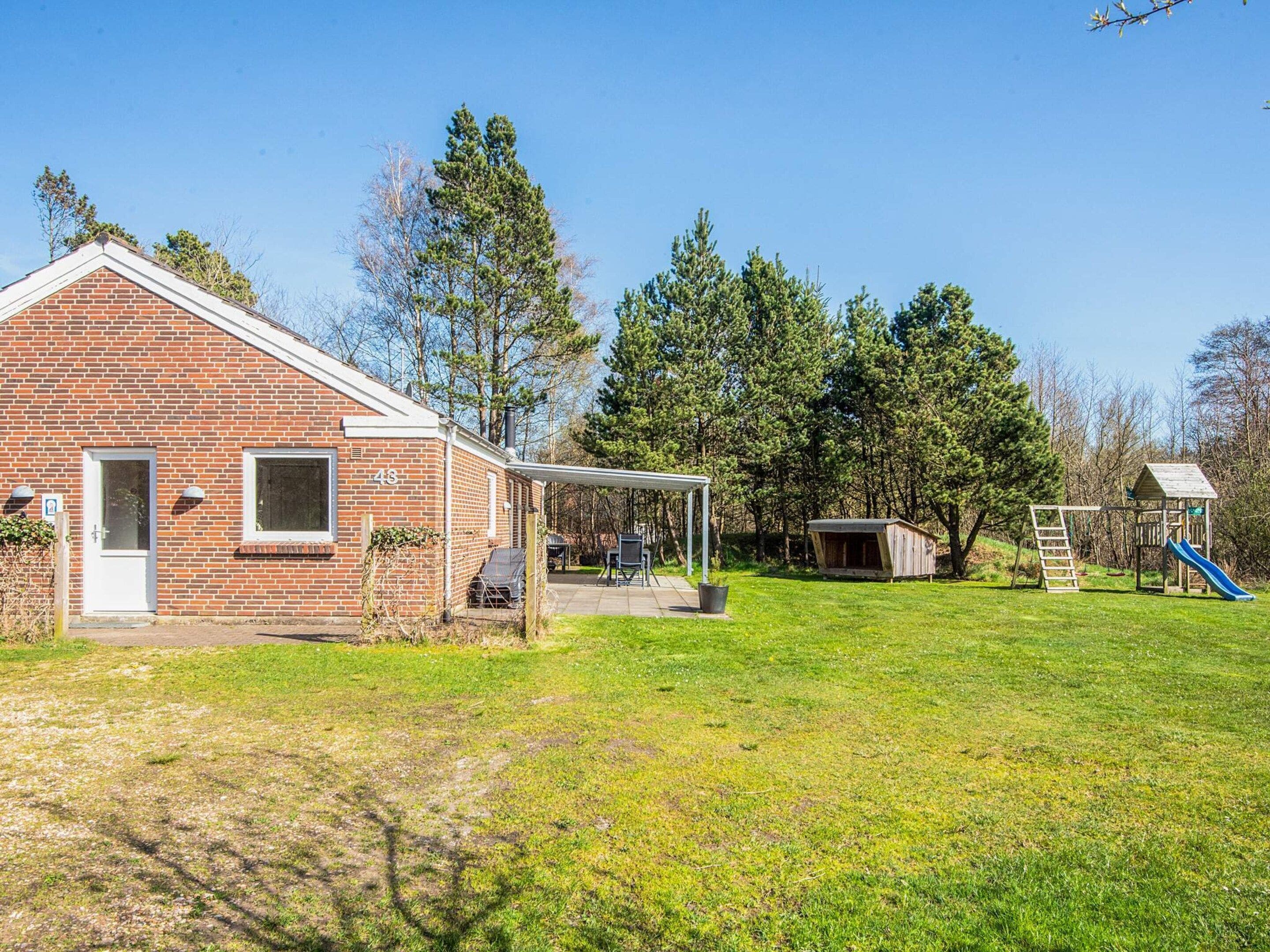 Une maison en briques avec un patio, un petit abri de jardin et un terrain de jeux équipé d’un toboggan.
