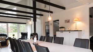 Un coin repas avec une longue table, des chaises noires, une nappe blanche et des luminaires suspendus. On y trouve également un canapé avec un coussin et une petite plante sur le c ôté.