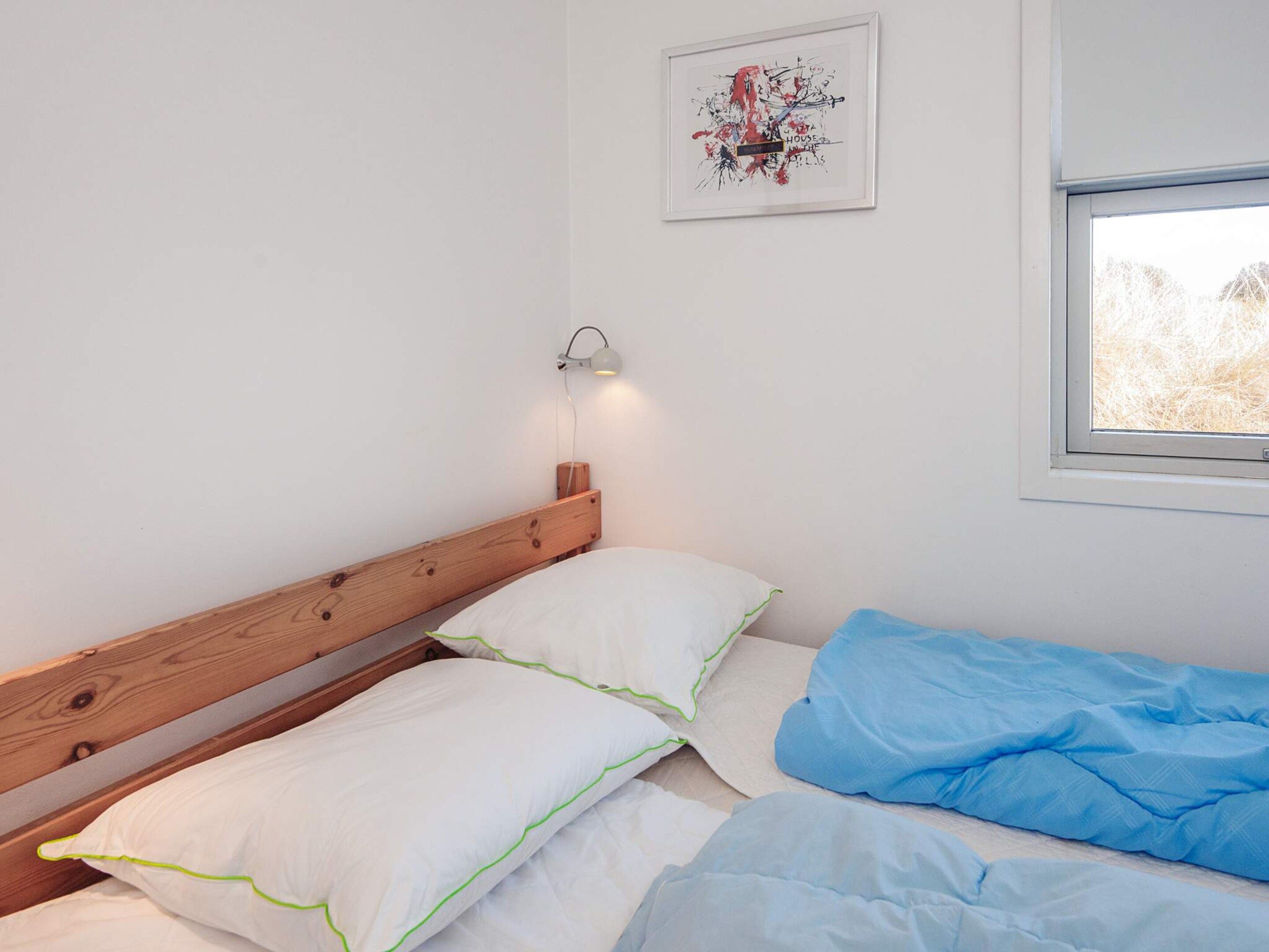 Une chambre avec une tête de lit en bois, un lit avec une literie blanche et bleue, une lampe fixée au mur et une fenêtre donnant sur des arbres.
