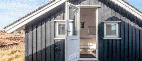 Una cabaña de madera oscura y pequeña con una puerta blanca abierta, mostrando un interior sencillo con una cama y un ventanal.